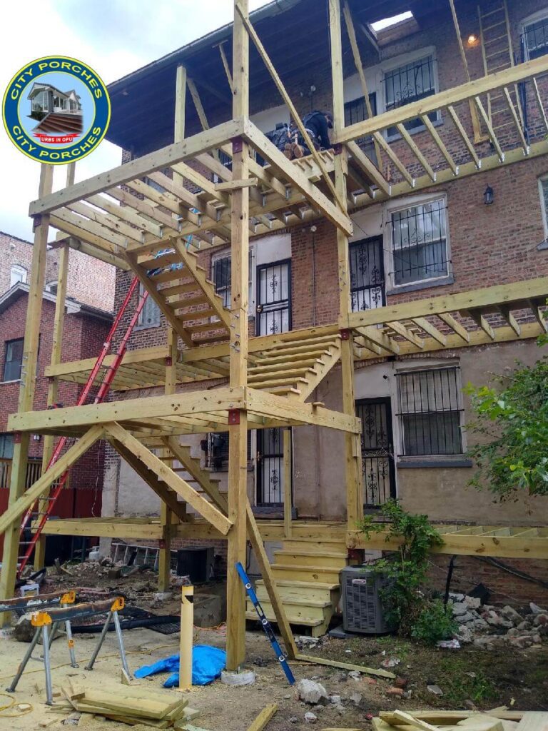 A construction scene with a new wooden staircase being built on the exterior of a brick apartment building. Tools and building materials are scattered around the site, indicating ongoing work. A worker can be seen standing on the upper level of the structure.