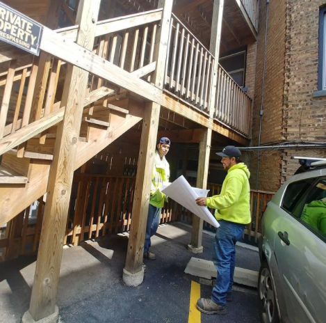 Two workers in high-visibility jackets discussing plans in front of a wooden staircase outside a brick building.