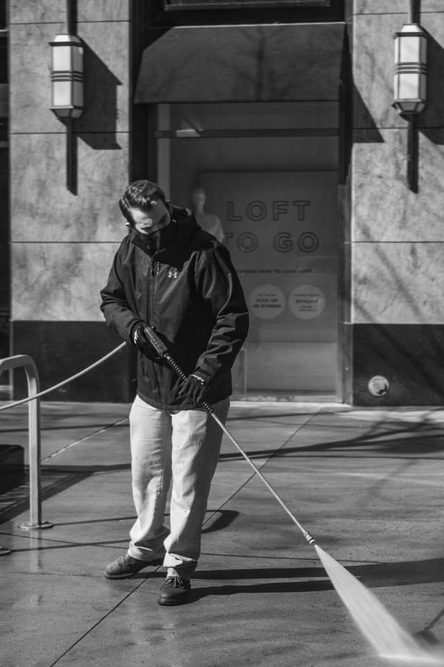 A person pressure washing a sidewalk in sunlight, outside a building with the signage "LOFT TO GO."