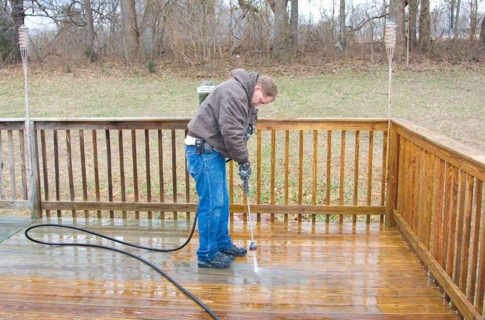 A man pressure washing a wooden deck with a railing, in a backyard setting.