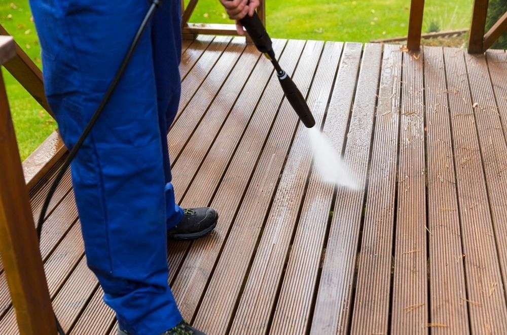 A person in blue coveralls pressure washing a wooden deck, with a focus on the high-pressure water jet cleaning the wood's surface, showing a clear difference between the cleaned and untreated areas.