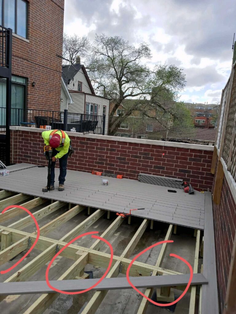Worker installing composite decking on a rooftop framed by brick walls with red marks on the wooden joists.- Porch Repair Chicago