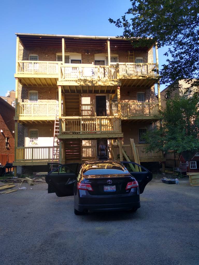 Rear view of a multi-story building with wooden porches, a parked car in the foreground.