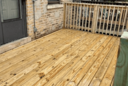 Close-up of a wooden deck floor with natural grain, next to a brick wall and a wooden railing.