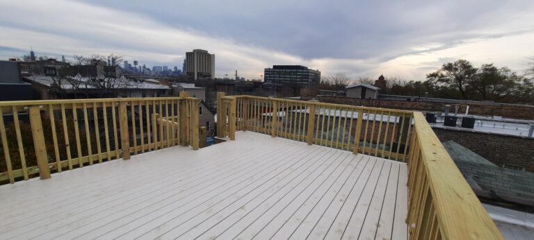 A spacious rooftop deck with fresh wooden flooring and railing, under an overcast sky with a view of urban buildings in the distance.