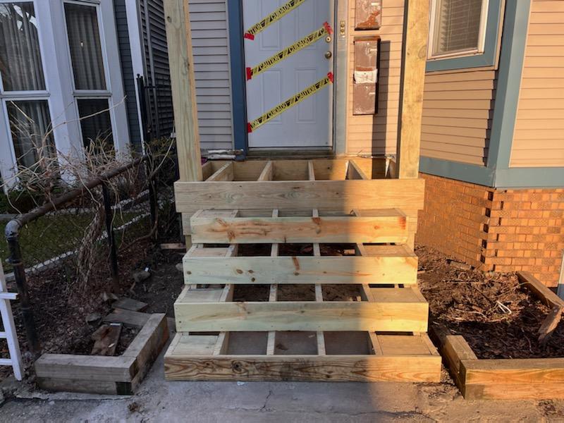 Newly constructed wooden porch steps with caution tape across the entrance and surrounding bare garden beds.- STAIR RISERS, multiporches