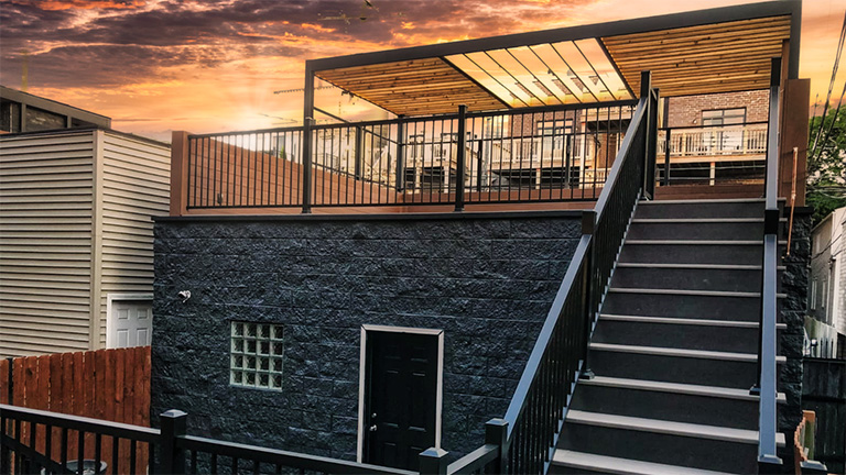 Sunset view of a modern terrace with glass cover, wooden ceiling, and metal railing above a dark stone facade with a black door and staircase.