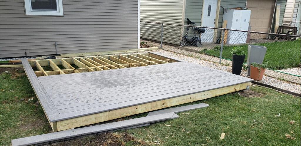 Partially constructed wooden deck with beam grey planking next to a grey house, with green lawn and fence in view.