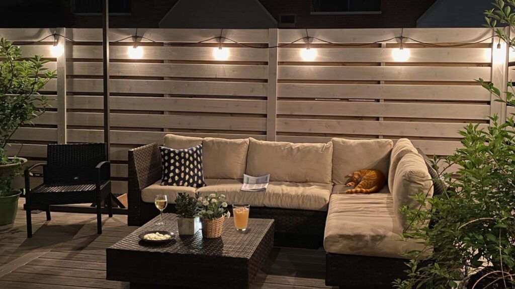 Outdoor evening setting with cozy sofa, chairs, and a coffee table, highlighted by warm string lights and lush plants.-Furniture Decking