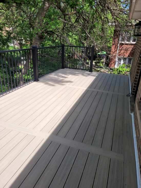 A beige composite deck with black railing, shaded by tree branches, in a residential backyard setting.