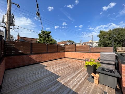 Spacious porch with deck with wooden flooring and privacy fencing, a grill, and potted plants under a blue sky.