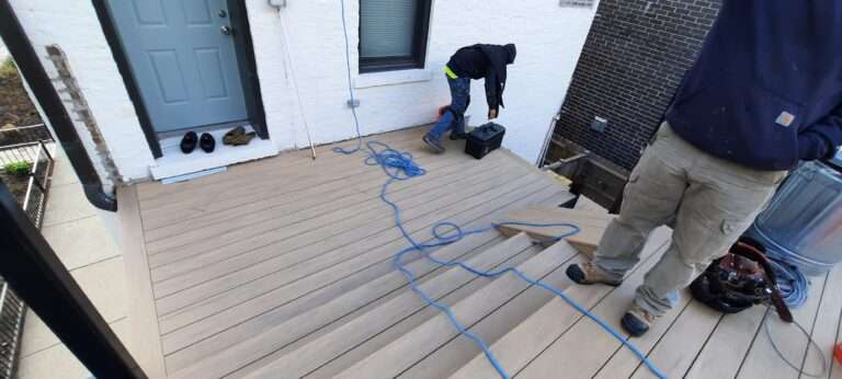 Porch construction worker bending over on a composite deck with tools, near a blue extension cord and a door with shoes beside it.