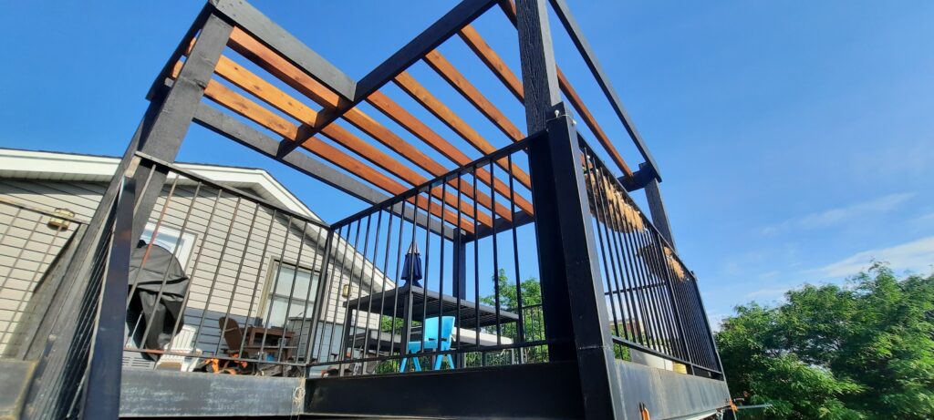 Low-angle view of a deck with black metal railings and a wooden pergola against a clear blue sky.-Pergola City
