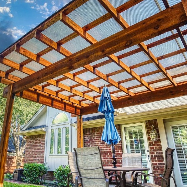 A porche covered area with a wooden pergola overhead, outdoor furniture, and a folded blue umbrella, next to a brick house with large windows.