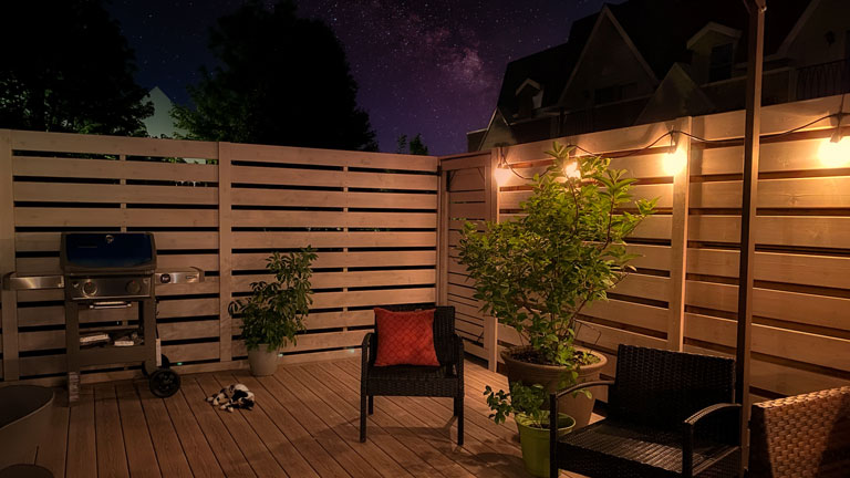 Cozy backyard deck at night, lit by string lights, with seating, a barbecue grill, and lush potted plants under a starry sky.