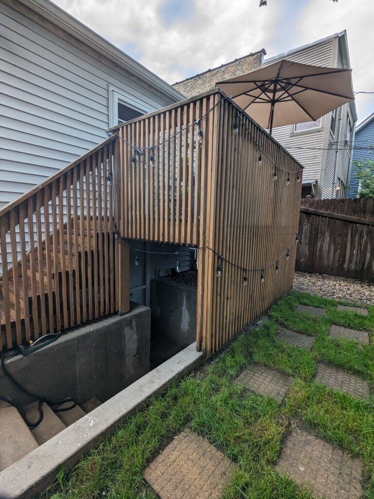 Wooden deck with vertical railing and an open umbrella, beside a concrete stairwell leading to a basement entrance.