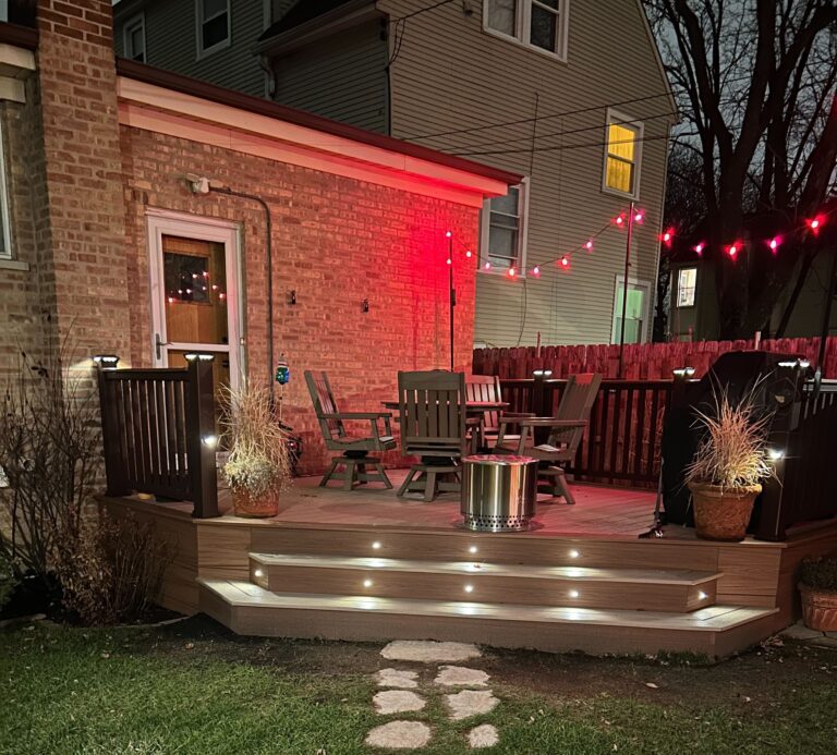 A cozy backyard deck at night illuminated with red string lights, featuring railing kits for deck, comfortable seating around a fire pit, and decorative plants adjacent to a brick house.-Trex Decking Cost