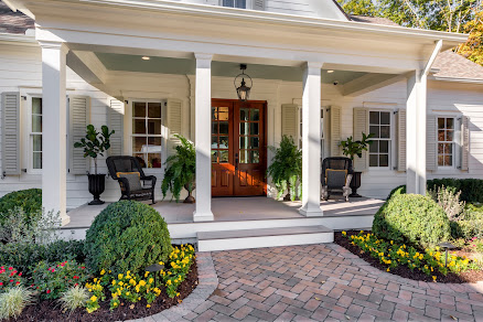 Spacious porch with white columns and a hanging lantern, framed by lush greenery and colorful flowers, leading to an elegant double door entry.