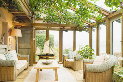 Bright and airy enclosed porch with a glass roof, lush greenery, and a set of wicker armchairs around a wooden coffee table.