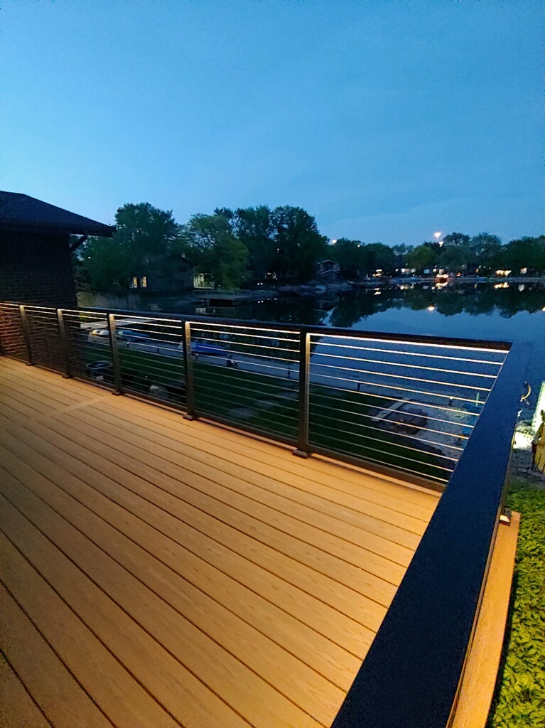Lakeside view from a wooden deck with stainless steel railings, evening lights illuminating the scene.