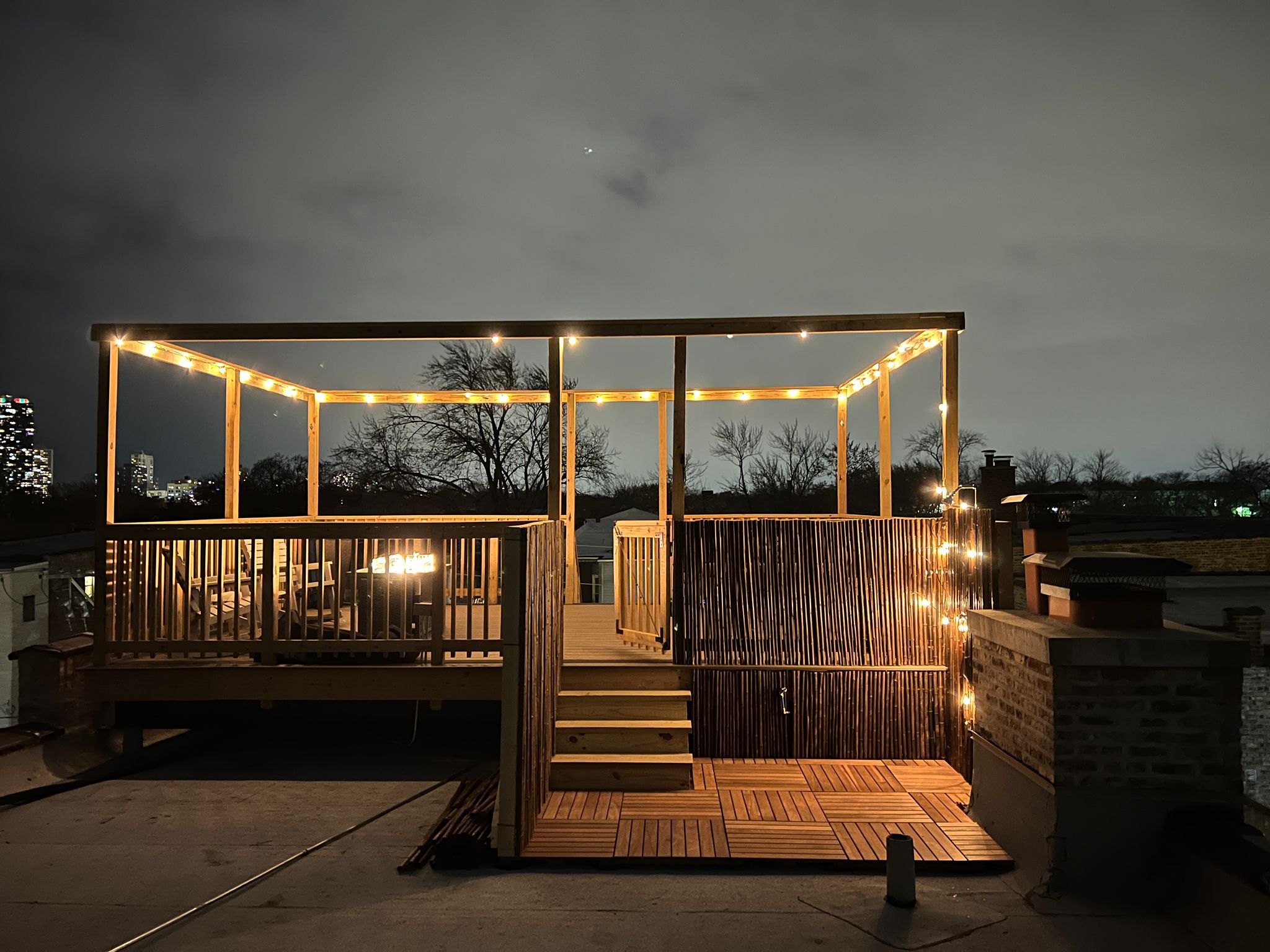 Rooftop deck with a wooden pergola, string lights, and a bamboo privacy screen, overlooking the city skyline at night.