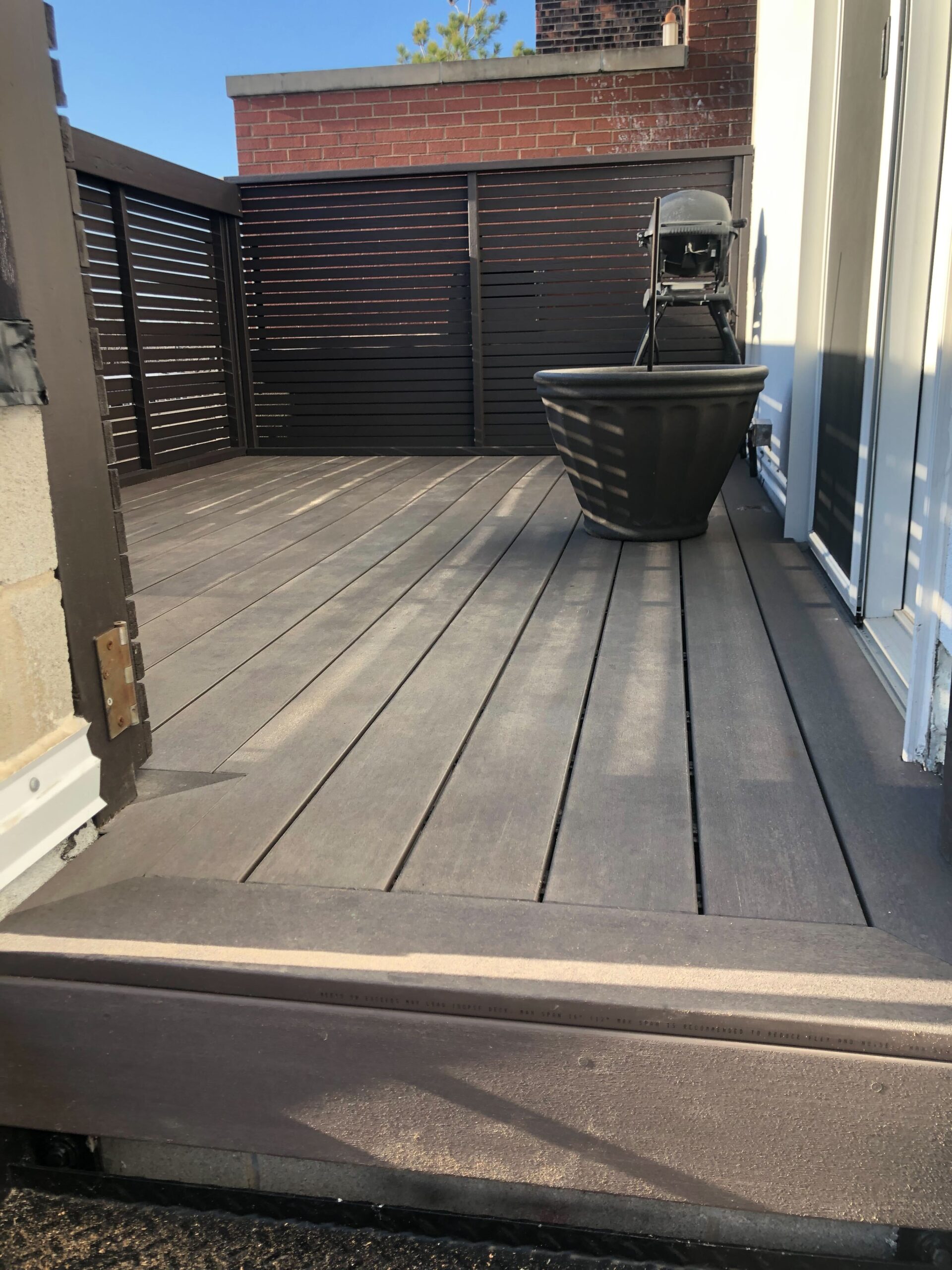 A modern outdoor deck with wooden flooring, a large planter, and a grill in the sunlight, surrounded by privacy slats.