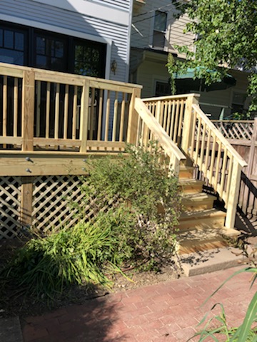 A light brown wooden deck with wooden railings and stairs leading down to a brick patio. The deck is made of wooden planks and has a lattice pattern underneath. The stairs have three steps and are made of wood. The patio is made of red bricks and has a brick path leading to the deck. There are some green plants growing around the base of the deck. The image is taken from a low angle, and the background is blurry.