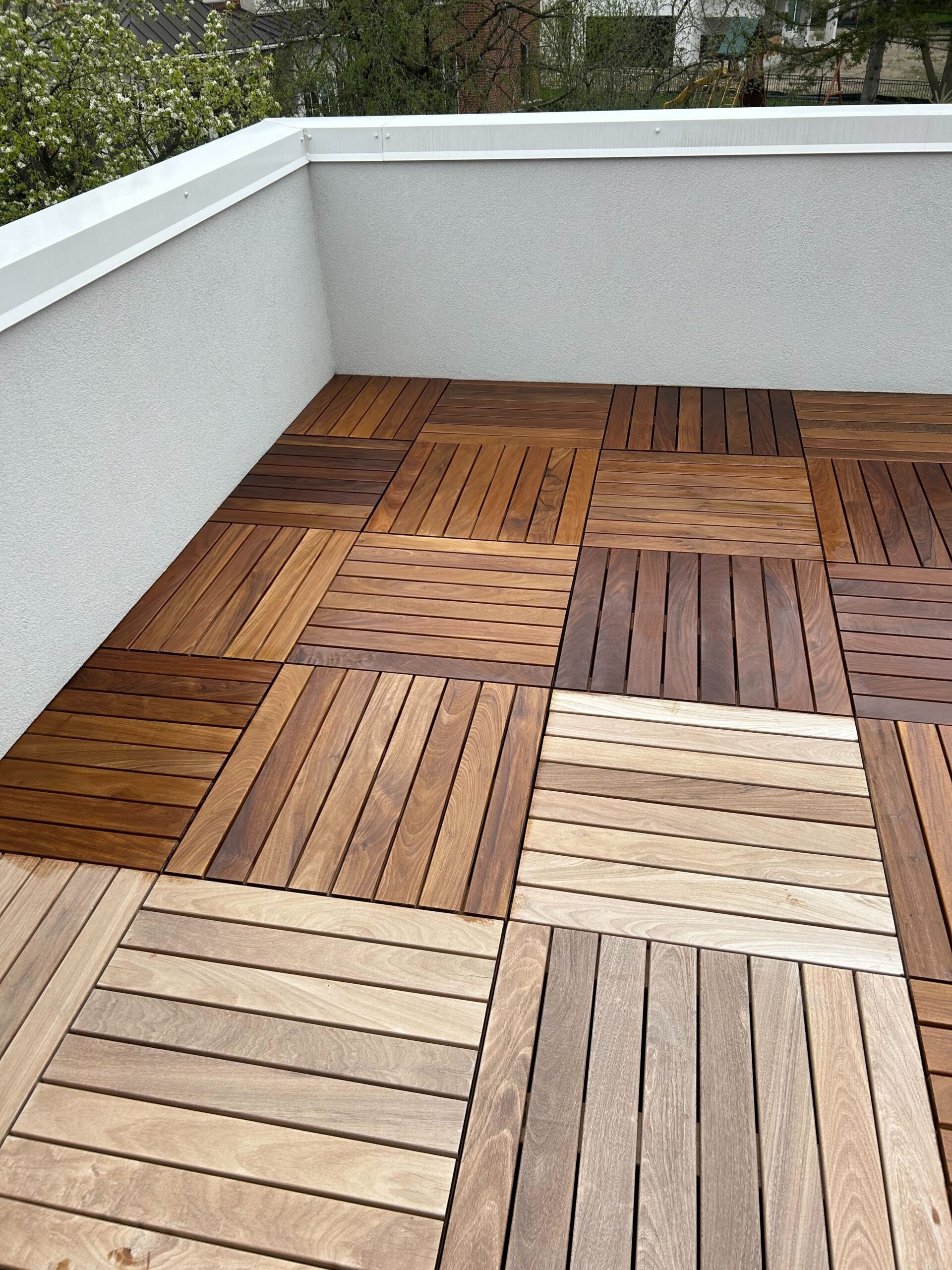 A rooftop terrace with wood tiles of different colors. The tiles are laid out in a checkerboard pattern. The terrace has a white wall around it. The image is taken from a high angle, and the background is blurry.