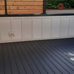 Grey deck flooring with a white paneled base of a wall and an outdoor electrical outlet.-Pergola City