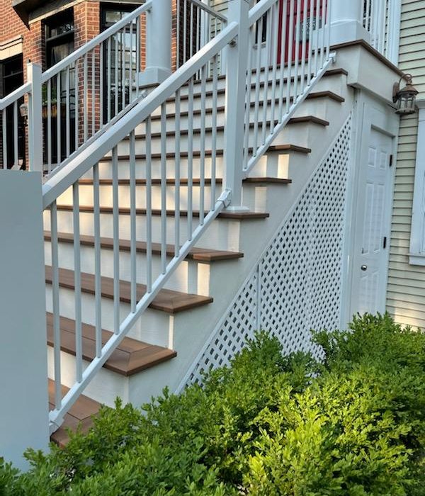 White exterior staircase with wooden steps, lattice side panels, and a railing, set against a house with green shrubbery.