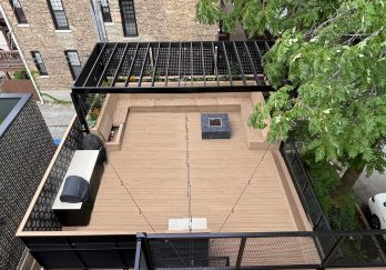 Overhead view of a rooftop deck with beige flooring, black railing, surrounded by trees and buildings