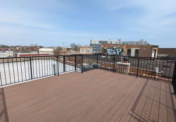 A spacious rooftop deck with brown composite decking and black metal railings, offering a wide view of the surrounding urban landscape with buildings in the distance.